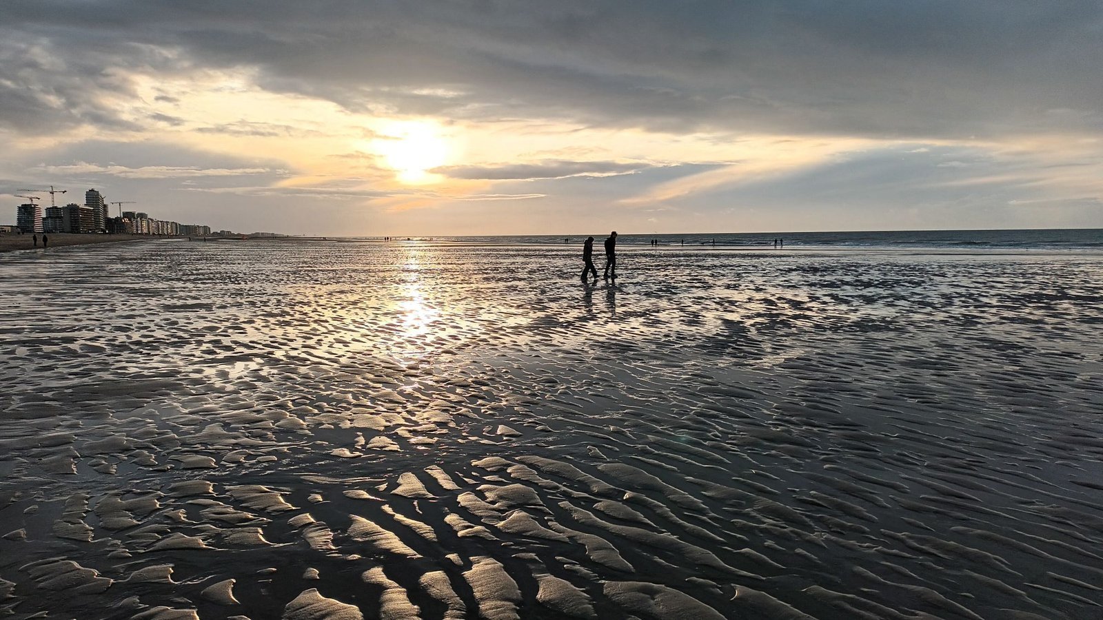 Two people walking across rippled wet sand at low tide under a golden, cloudy sunset on the coast
